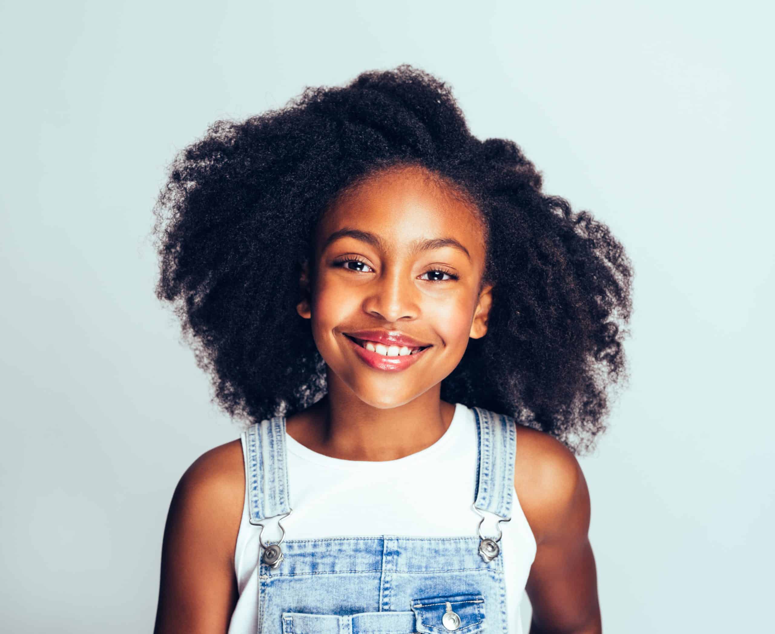 Smiling young African girl with long curly hair wearing dungarees standing happily against a gray background