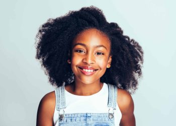 Smiling young African girl with long curly hair wearing dungarees standing happily against a gray background